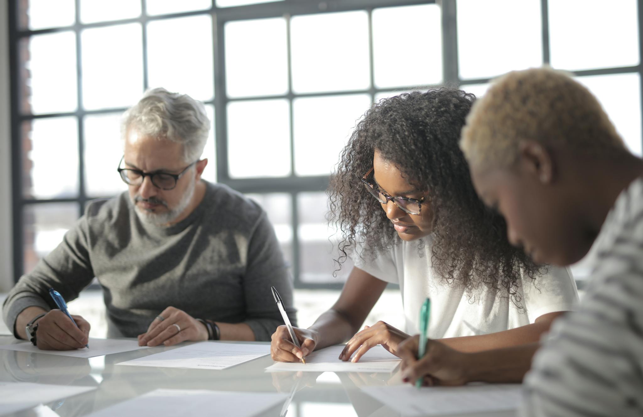 Serious multiethnic colleagues in casual wear writing on papers with pen during teamwork while sitting at table in light office behind fenced window